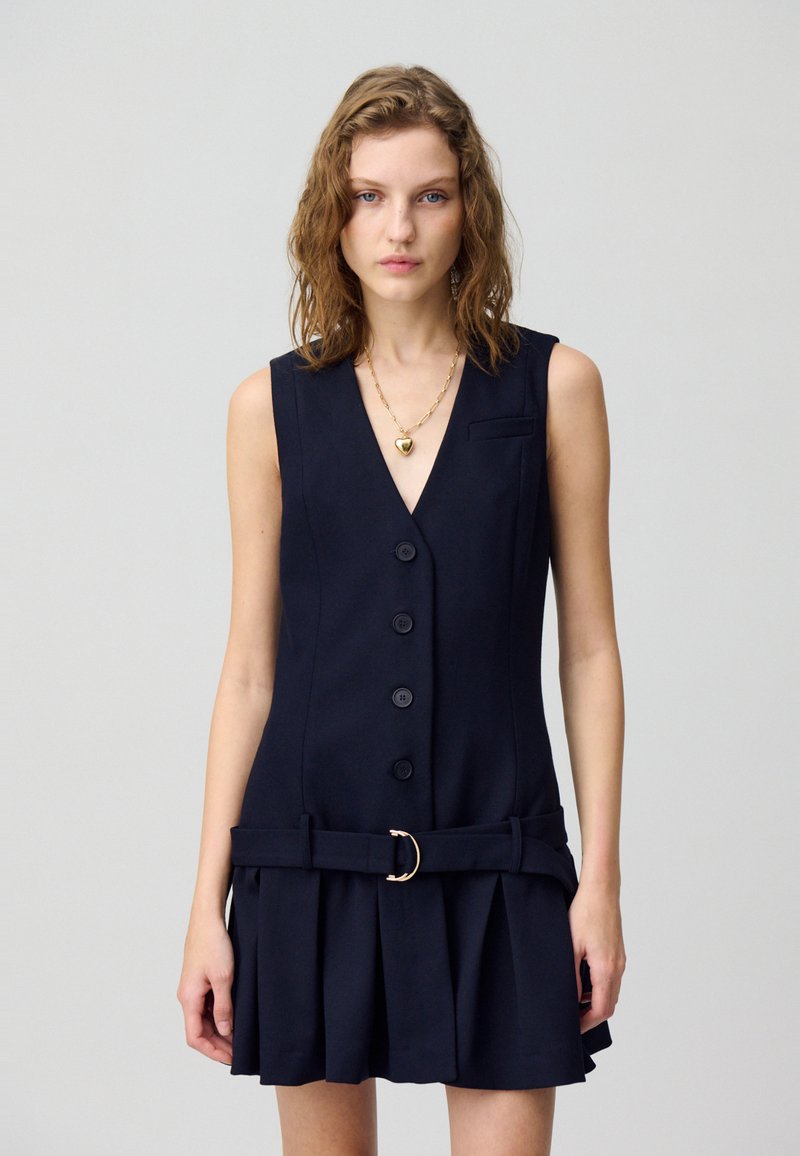 Robe sans manches bleu marine au design ajusté, avec un col en V, des boutons sur le devant et une taille ceinturée avec une boucle circulaire dorée. Détails de jupe plissée.