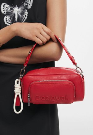 Bolso de mano Desigual de cuero rojo con un bolsillo delantero con cremallera y llavero de cuerda blanca, sostenido por una persona que lleva una camiseta negra con un estampado de mariposas.