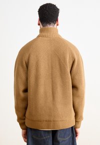 Brun ribbad stickad tröja med hög krage och sömdetaljer på baksidan; har en avslappnad passform och ärmar. Bärs över mörka jeans.