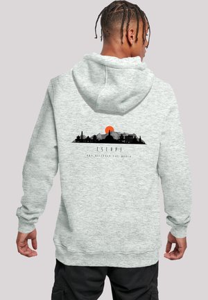 Person with braided hair wearing light gray hoodie with black city skyline and orange sun graphic and text "Escape, not discover the world" on back.
