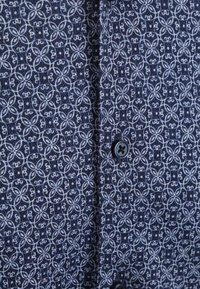 Blauwe shirt met een geometrisch patroon en lichtblauwe accenten. Zwarte knop in het midden. De stof lijkt textuur te hebben.