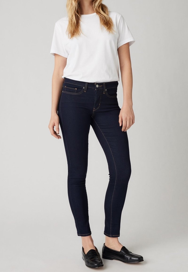 Femme portant un t-shirt blanc à manches courtes, un jean skinny bleu foncé et des mocassins noirs, debout devant un fond uni gris clair.