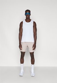 Λευκή αμάνικη μπλούζα με μικρή λογότυπο, μπεζ σορτς, λευκά αθλητικά παπούτσια και μεγάλα μπλε γυαλιά ηλίου. Μαλακό ύφασμα και αθλητική εφαρμογή.