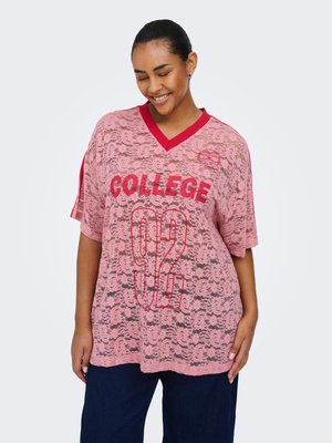 Smiling woman wears pink lace oversized top with red "COLLEGE 92" text and blue pants, standing against plain light background.