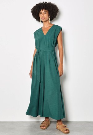 Donna con capelli ricci che indossa orecchini d'oro, un abito maxi verde senza maniche con cintura e sandali beige, in piedi contro un muro bianco.