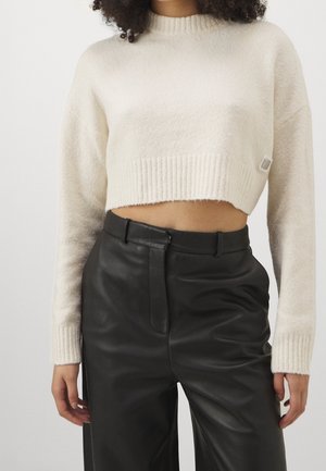 Frau trägt einen kurzen weißen Strickpullover mit langen Ärmeln und hoch taillierte schwarze Lederhose, steht vor schlichtem Hintergrund.