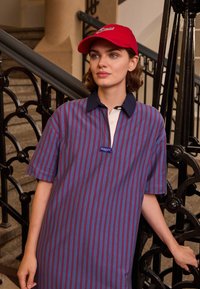 Vestido de camisa listrado em azul e vermelho com um colarinho branco e o logótipo GANT. Combinado com uma boné vermelho, tecido texturizado, corte solto, mangas curtas.