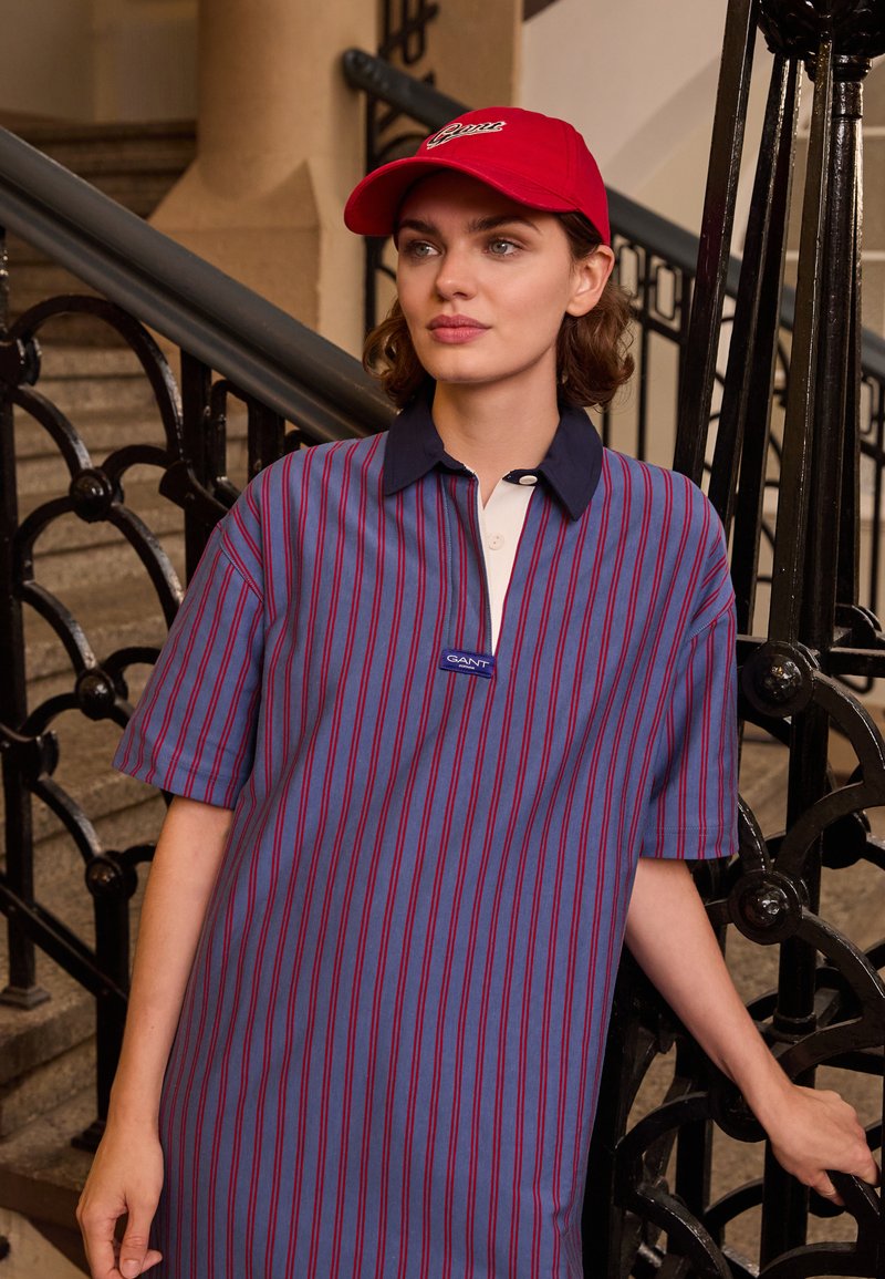 Vestido de camisa listrado em azul e vermelho com um colarinho branco e o logótipo GANT. Combinado com uma boné vermelho, tecido texturizado, corte solto, mangas curtas.