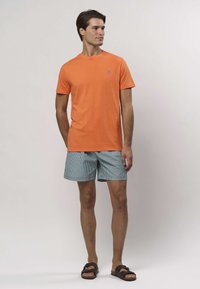 Svetlooranžové bavlnené tričko s malým modrým logom. Kombinované s modro-bielymi pruhovanými šortkami. Model má na nohách hnedé sandále.