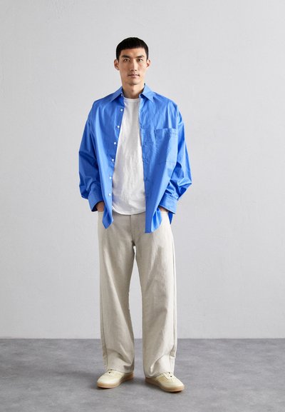 Completo maschile composto da una camicia azzurra brillante con bottoni, una t-shirt bianca, pantaloni beige larghi e scarpe da ginnastica chiare su un pavimento grigio.