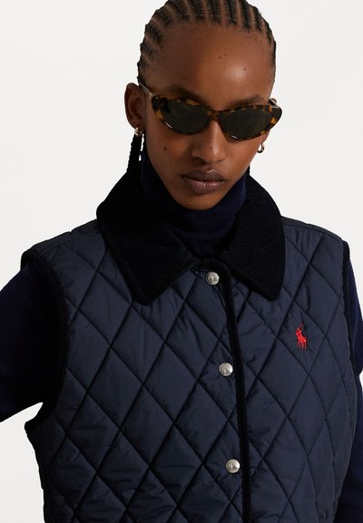 Marineblauwe gewatteerde vest met een corduroy kraag, zilveren drukknoopjes en een klein rood logo op de borst. Draag het over een donkere coltrui.