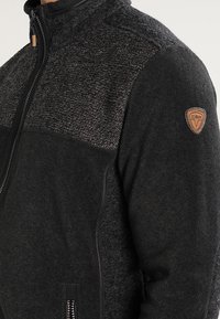 Schwarze Fleecejacke mit strukturiertem Oberteil, Reißverschluss, Seitentaschen und einem braunen Logoaufnäher an der linken Schulter.