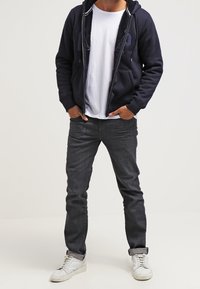 Sweat à capuche zippé marine avec doublure en polaire, t-shirt blanc à col rond, jean en denim foncé et baskets de couleur claire. Comprend des poches avant.