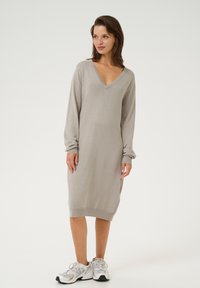 Robe longue gris clair à manches longues avec un décolleté en V, texture en tricot douce et détails de poignets côtelés, associée à des baskets blanches et grises.