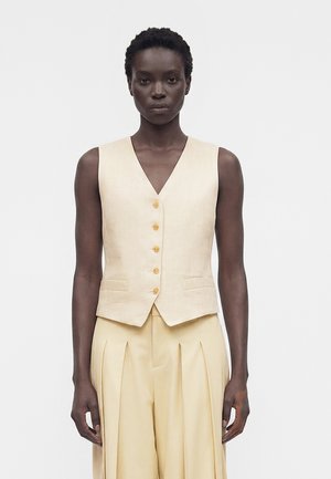 Kobieta o krótkich włosach, ubrana w beżową, bezrękawnikową kamizelkę na guziki oraz plisowane, z wysokim stanem spodnie, na jednolitym tle.