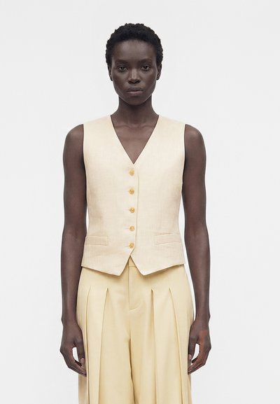Femme aux cheveux courts portant un gilet beige sans manches boutonné et un pantalon plissé taille haute sur un fond uni.