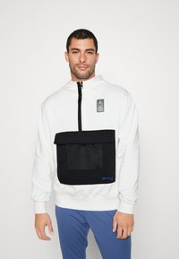 Hvid hoodie med en sort netlomme og klap, prydet med et logo-applikation og lynlås-lukning. Blå tekstdetalje på lommen fremhæver designet.