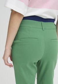 Pantalon vert à texture lisse, conçu avec une taille ajustée et deux poches arrière ; la ceinture présente un accent de couleur bleu marine.