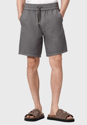 Mężczyzna noszący szare szorty z elastyczną talią i sznurkiem oraz brązowe sandały na wsuwki, ręce w kieszeniach, stojący na prostym jasnym tle.