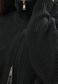 Svart stickad zip-up tröja med hög krage, med ribbstruktur och silverfärgad dragkedjedetalj. Närbild av tyget och stängningen.
