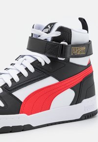 Svart och vit högskurna sneakers med en röd accent. Har en texturerad läderfinish, vita snören och ett kardborreband med guldtryck.