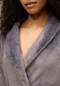 Pilkas, minkštas, pūkuotas chalatas su šaliku apykakle, turintis lygią tekstūrą ir laisvą siluetą, išryškinantis gilų iškirptę ir minimalias siūlių detales.