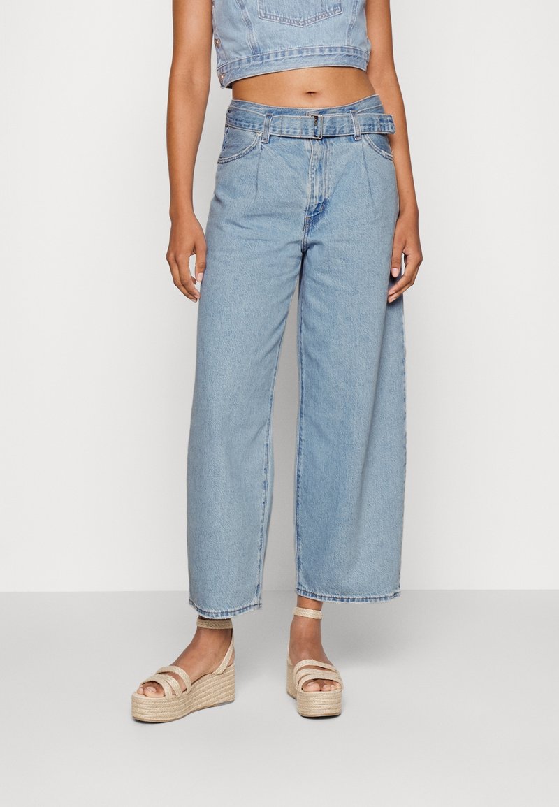 Femme portant un jean taille haute bleu clair à jambes larges avec une ceinture, associé à un top court en denim et des sandales compensées beiges.