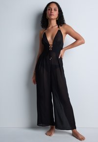 Femme aux cheveux bouclés portant une combinaison noire sans manches en tissu transparent avec des jambes larges, se tenant pieds nus contre un mur clair.