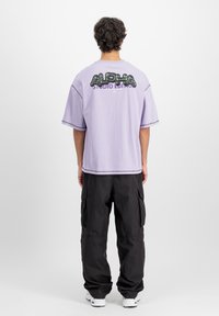 Tricou oversized violet cu un imprimeu grafic negru pe spate, asociat cu pantaloni cargo negri și adidași albi.