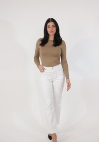 Femme aux longs cheveux bruns portant un haut beige ajusté à manches longues, un jean blanc et des chaussures noires pointues, debout devant un fond blanc.