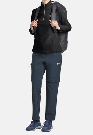 Czarna bluza z polaru z połową zamka, granatowe elastyczne spodnie z zamkami błyskawicznymi oraz szary plecak. Niebieskie buty trekkingowe. Funkcjonalny strój outdoorowy.