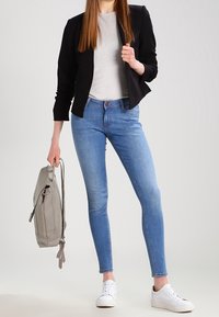 Černý zkrácený blazer, světle šedé tričko, modré úzké džíny a bílé tenisky. Světle šedý batoh je držen v levé ruce.