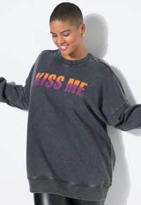Sudadera de gran tamaño en gris oscuro con dobladillo y puños acanalados. Presenta un texto degradado "KISS ME" en naranja y púrpura en la parte frontal.