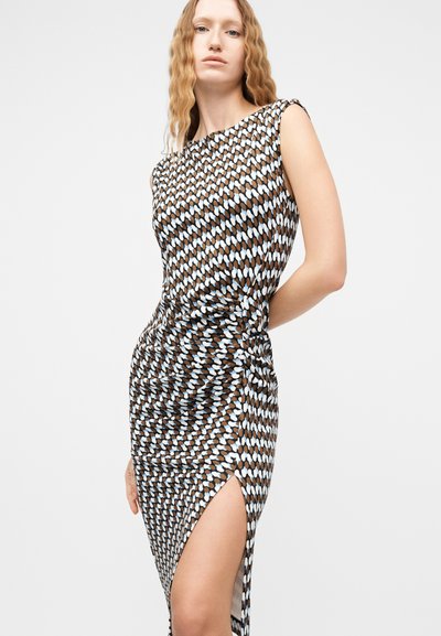 Γυναίκα με μακριά κυματιστά μαλλιά που φοράει αμάνικο φόρεμα με σχέδια και υψηλή σχισμή, στέκεται μπροστά σε απλό φωτεινό φόντο.