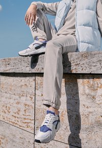 Des chaussures de sport blanches et violettes avec des accents en mesh reposent sur un mur en pierre. La personne porte un pantalon de jogging gris et un gilet rembourré bleu clair.