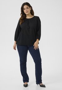 Zwarte blouse met stippen, pofmouwen, ronde hals en een relaxte pasvorm, gecombineerd met donkerblauwe rechttoe-rechtaan jeans en zwarte puntige hakken.