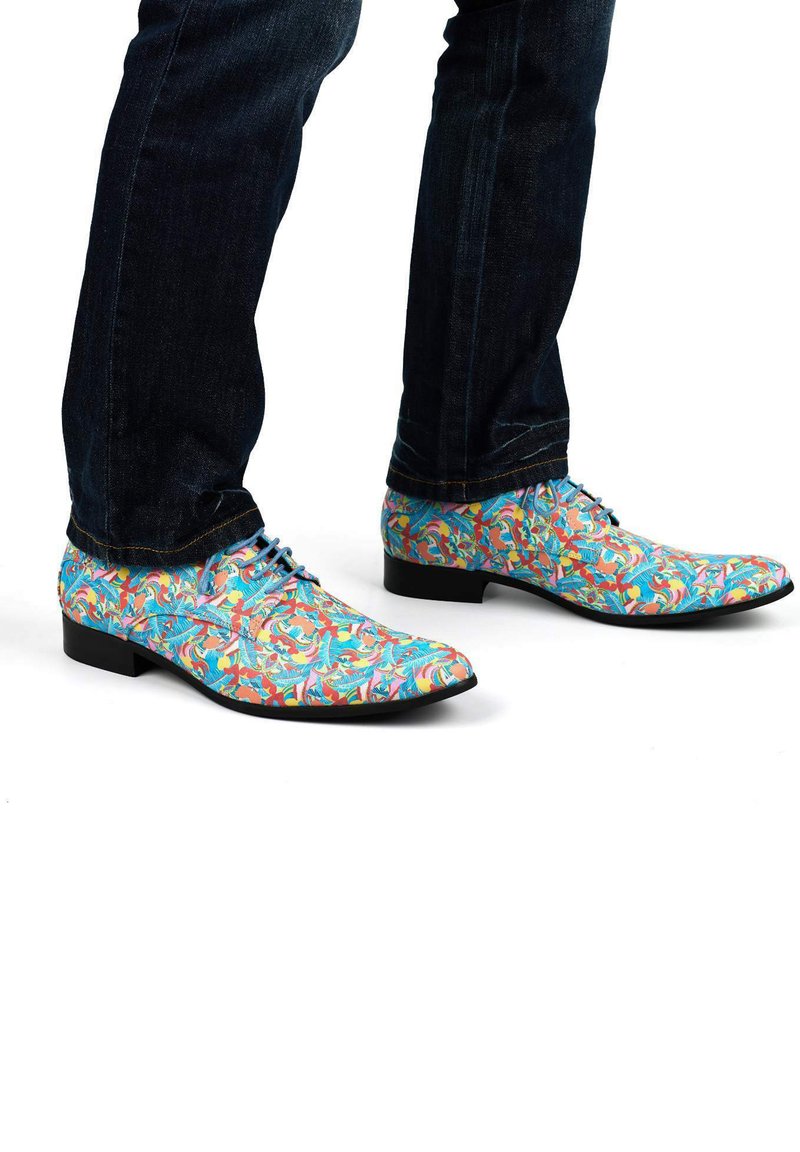 Chaussures habillées colorées avec une base bleue, ornées de motifs floraux complexes en rose, orange et vert, associées à un jean foncé.