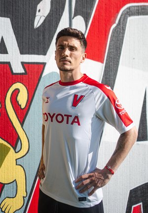 Wit voetbalshirt met rode accenten en het "TOYOTA"-logo op de voorkant. Korte mouwen, ronde hals, gestructureerde stof. Op de achtergrond een kleurrijk muurschildering.