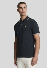 Svart polo-skjorte med hvit krage og mansjetttrim, med en gul brodert logo på brystet. Laget av glatt bomullsstoff.