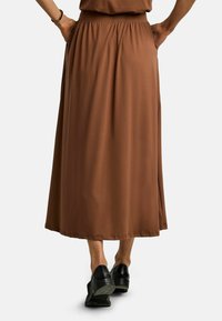 Sieviete valkā brūnu pie potītēm garu svārku ar elastīgu jostasvietu, melnus zemas papēžus apavus, abas rokas kabatās, redzama no muguras.