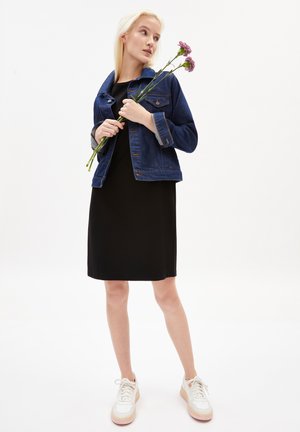 Dunkelblaue Jeansjacke mit goldenen Nähten, schwarzes Kleid und weiße Sneakers mit beigen Akzenten. Modell hält pinke Blumen.