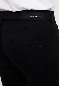 Jeans de denim negro con una textura suave. Las características incluyen un parche de cuero REPLAY en la cintura y un bolsillo trasero con un sutil detalle de logo.