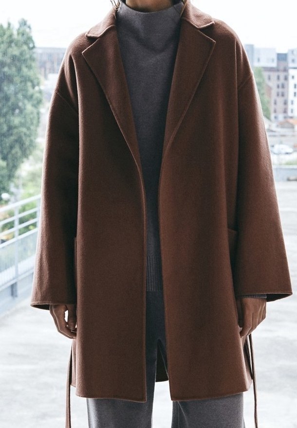Cappotto in lana marrone con un ampio colletto, vestibilità larga e maniche lunghe. Presenta tasche laterali e un design a taglio dritto. La trama del tessuto è visibile.