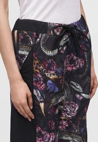 Κοντινή λήψη παντελονιών με floral και ζωικό σχέδιο και μαύρο κορδόνι, φορεμένα με λευκό τοπ, δείχνει ένα χέρι που ακουμπάει στο πλάι.