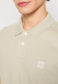 Polo beige en tricot texturé, doté d'une patte de boutonnage à trois boutons, d'un col classique et d'un logo embossé discret sur la poitrine.
