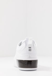 Vit idrottssko med en slät syntetisk ovandel, förbättrad dämpning vid hälen och svarta gummidetaljer på sulan. Nike-logotyp ingår.