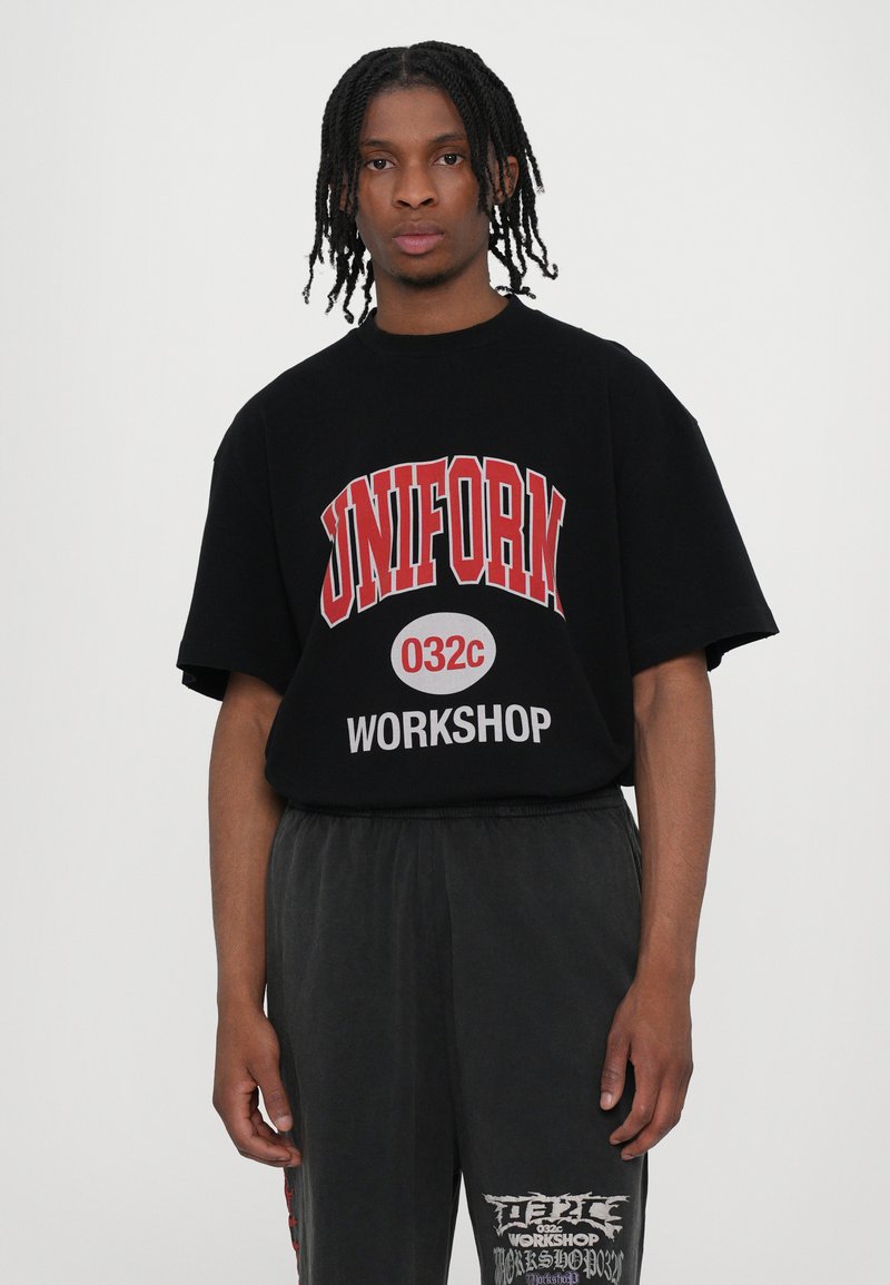 Camiseta negra de gran tamaño con la inscripción "UNIFORM 032c WORKSHOP" en letras rojas y blancas, combinada con pantalones grises que presentan acentos gráficos.