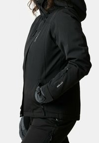Personne portant une veste noire à capuche avec poches zippées et un pantalon noir assorti, mains dans les poches, sur un fond blanc uni.