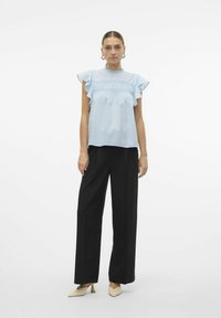 Blusa azzurro chiaro a maniche corte con spalline arricciate e dettagli smock, abbinata a pantaloni larghi neri e scarpe beige a punta.