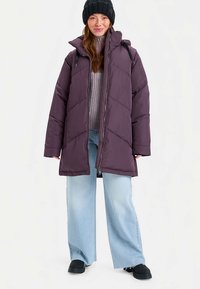 Lila Steppmantel mit Kapuze, mit diagonalem Steppmuster und Reißverschlüssen. Getragen über einem leichten Pullover und Jeans mit weitem Bein. Schwarze Mütze und Schuhe.